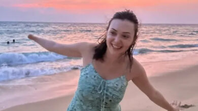 Seher Başaran, sahilde sağ kolunu yukarı ve sol kolunu aşağı doğru tutarak, ayakta poz veriyor.