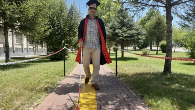 Görme engelli Emek Çağatay Aksöz, kep, cüppe giyip, siyah bir gözlük takmış ve beyaz bastonu görme engelli yolunda kullanarak yürüyor.