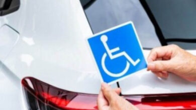 Beyaz renkli bir otomobil ve engelli bireylere ait özel alanlardan yararlanması için tasarlanmış mavi zemin üzerinde tekerlekli sandalye kullanan biri var.