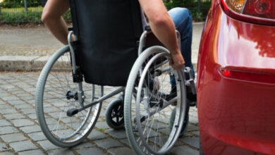 Tekerlekli sandalye kullanan engelli birey, kırmızı renkli bir otomobilin sürücü koltuğuna oturmak için araca yaklaşıyor.