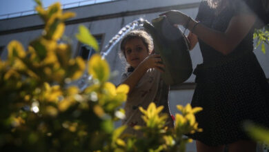 Bir kız çocuğu, yetişkin bir kadının desteğiyle çiçeklere su döküyor.