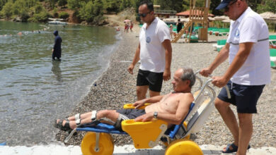 Tekerlekli sandalye kullanan engelli birey, denize girmek için özel tasarlanmış bir tekerlekli sandalye üzerinde ve denize doğru giden engelli rampası ile görevli iki kişi sandalyeyi sürüyor.