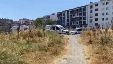 Görselde boş bir alan ve çalı boyutlarında otlar, ileri bir yerde ambulans ve polis araçları var.