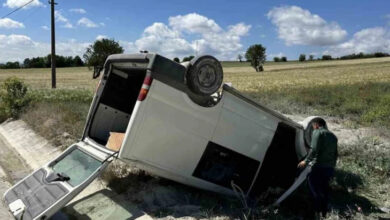 Beyaz renkli bir minibüs yol kenarında ters dönmüş şekilde bagaj kapağı ve sürücü koltuğu kapısı açık şekilde duruyor.
