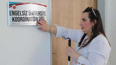 Fotoğrafta görme engelli birey, bir elinde beyaz bastonu ve diğer eli ile duvara asılı tabeladaki braile alfabesi ve üstünde büyük puntolarla "Engelsiz Üniversite Koordinatörlüğü" yazısı var.