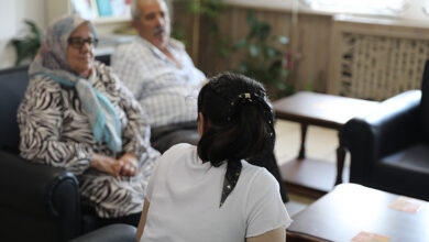 Zihinsel engelli Ayşe, kameraya sırtı dönük şekilde koltukta oturuyor. Fotoğrafın sol köşesinde yan yana koruyucu aile oturuyor.
