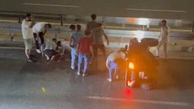 Fotoğraf yukarıdan yatay bir yerden çekilmiş. Dört tekerlekli arazi aracı yan yatmış ve yerde yatan bir kişinin etrafına kalabalık toplanmış.