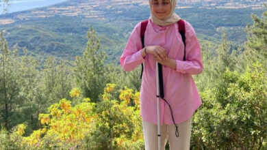 Fotoğrafta Berra Yami, ormanlık bir alanda, arka planda geniş bir manzara ve deniz görülen bir yerde duruyor ve kameray bakarak poz veriyor. Yami, pembe bir tunik giymiş, krem bir başörtüsü var. Ayrıca, sırtında kırmızı bir sırt çantası taşıyor. Elinde bir beyaz baston tutuyor.