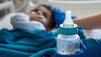 Fotoğrafta, yatakta uzanmış küçük bir bebek ve sağlık personeli kıyafeti içerisinde biri belinden aşağısı gözükecek şekilde başında bekliyor. Sağlık personelinin elinde bebeğin beslenmesi için biberon var.