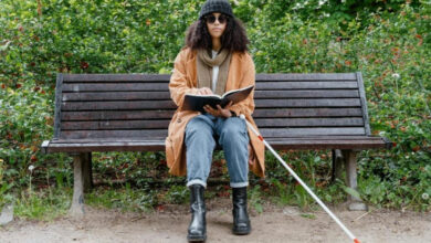Görme engelli birey bankta oturuyor ve yanında beyaz baston, elinde ise braile alfabesi ile yazılmış kitabı parmak ucuyla dokunarak okuyor.