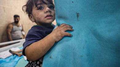 Küçük bir çocuk annesinin kucağında, omzunun üstünden arkaya doğru bakıyor. Çocuğun arkasında, bir sedyenin üzerinde başka bir çocuğun ayağı gözüküyor. Sedyenin diğer tarafında ise beyaz atlet giymiş yetişkin bir erkek var.