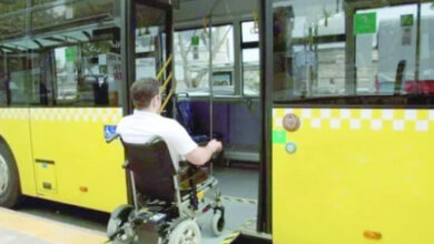 Akülü tekerlekli sandalye kullanan engelli birey, sarı renkli belediye otobüsüne engelli rampası ile biniyor.