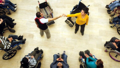 Tekerlekli sandalye kullanıcısı engelli bireyler çember oluşturacak şekilde yan yana duruyorlar. çemberin ortasında boş iki tekerlekli sandalye var. Tekerlekli sandalye önünde ise bir erkek ile kadın ayakta duruyor.