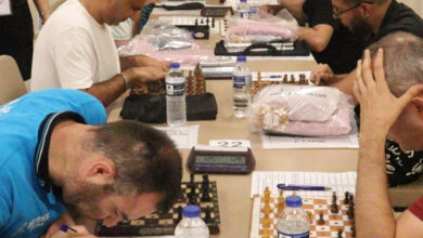 Görme engelli bireyler karşılıklı masa çevresinde oturmuşlar. Masa üzerinde satranç takımları ve not kağıtları var.