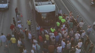 Yolun ortasında duran rayların üstünde tramvay vagonları var. Tramvayın ön bölgesinde 3 polis memuru ve çok sayıda insan kalabalığı olduğu görülüyor.