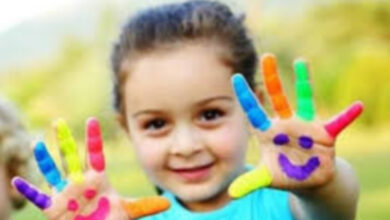 Fotoğrafta bir çocuğun her iki el parmakları farklı renklere boyanmış ve iki avucuna gülen surat resmi çizilmiş.