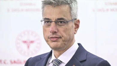 Sağlık Bakanı Kemal Memişoğlu, basın mensuplarını sorularını yanıtlıyor. Sağlık Bakanlığı’nın arkasında ise beyaz bir duvarın üzerine yerleştirilmiş “Sağlık Bakanlığı” yazıları olan birçok amblem yapıştırılmış.