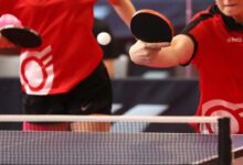 Fotoğrafta iki kişi var. Masa tenisi oyunu sırasında bir kişi vuruş yapıvor ve vuruş yapan kişinin yüzünün alt bölümü, diğer sporcunun belden aşağı fotoğraf içinde yer alıyor.