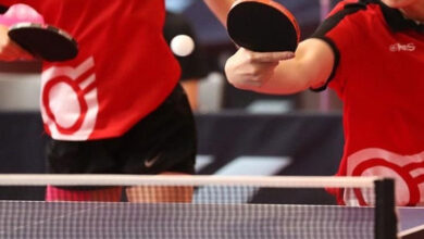 Fotoğrafta iki kişi var. Masa tenisi oyunu sırasında bir kişi vuruş yapıvor ve vuruş yapan kişinin yüzünün alt bölümü, diğer sporcunun belden aşağı fotoğraf içinde yer alıyor.