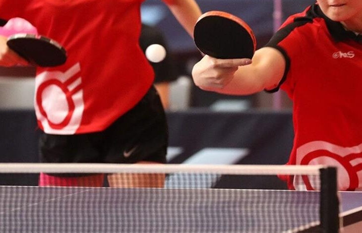 Fotoğrafta iki kişi var. Masa tenisi oyunu sırasında bir kişi vuruş yapıvor ve vuruş yapan kişinin yüzünün alt bölümü, diğer sporcunun belden aşağı fotoğraf içinde yer alıyor.