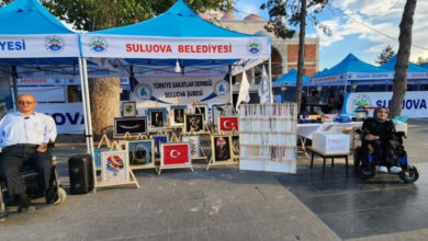Fotoğrafta, akülü tekerlekli sandalye kullanıcısı iki engelli birey, el işi ürünlerin satışının yapıldığı standın başında duruyor. El işi ürünlerin satıldığı yerde “Türkiye Sakatlar Derneği Suluova Şubesi” yazısı var.