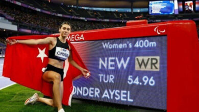 Paralimpik atletizm sporcusu Aysel Önder, koşu süresini gösteren sayacın önünde Türk bayrağını boynuna asarak poz veriyor.