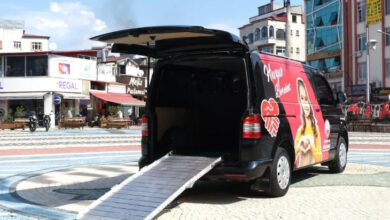 Fotoğrafta, siyah bir minibüsün arka bagaj kapısı açık ve kapının olduğu yerden zemine doğru engelli rampası açık bir şekilde duruyor.