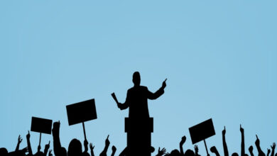 Görselde siyah renkli insan figürleri var. İnsan figürlerinden bir tanesi diğer kişileri karşısına alarak bir kürsünün arkasından konuşuyor. Diğer kişilerde kollarını kaldırarak ve kalabalık bir şekilde durarak destek veriyorlar.