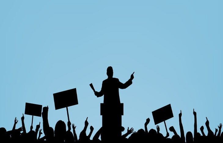 Görselde siyah renkli insan figürleri var. İnsan figürlerinden bir tanesi diğer kişileri karşısına alarak bir kürsünün arkasından konuşuyor. Diğer kişilerde kollarını kaldırarak ve kalabalık bir şekilde durarak destek veriyorlar.