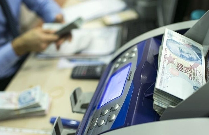 Para sayma makinesinin üzerinde 100 TL'lik banknotlar var. Fotoğrafta, flu bir şekilde hesap makinesi, 100 TL'lik banknotlar, bilgisayar ekranı ve klavyesi, masanın üzerinde duruyor. Aynı zamanda yüzü gözükmeyen bir kişi elindeki paraları sayıyor.