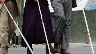 Beyaz baston ile caddede karşıdan karşıya geçmeye çalışan 3 görme engelli birey görselde var. Görme engelli bireylerin belden yukarısı görselde yer almıyor. Görseldeki kişilerden ikisi pantolon giydiği için erkek, bir kişi ise etek giydiği için kadın olduğu tahmin edilebilir.