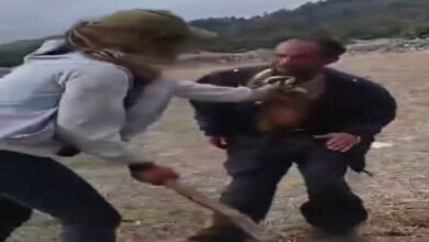 Fotoğrafta, engelli bir çobanı, yeşil bir alanda elinde sopa tutan bir kadın yakasından tutuyor.