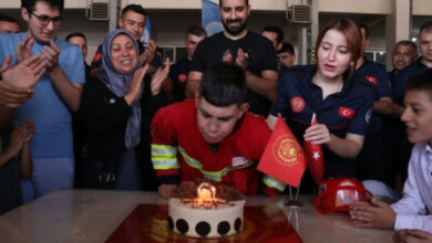 Fotoğrafta, bir masanın üzerinde yaş pasta var. Yaş pastanın üzerinde yanan mumlara doğru bir kişi eğilerek üflüyor. Arka taraflarında ise farklı yaşlarda birçok kişi var. Arka taraftaki kişilerden bazıları itfaiye üniforması giymiş.