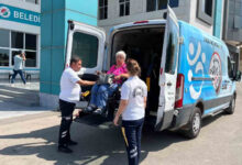 Akülü tekerlekli sandalye kullanıcısı engelli birey, bir minibüsün arkasına özel olarak yapılmış engelli asansörü ile araçtan aküllü sandalye üzerinde indiriliyor. Engelli bireyin yanında refakat ile belediye personelleri var.