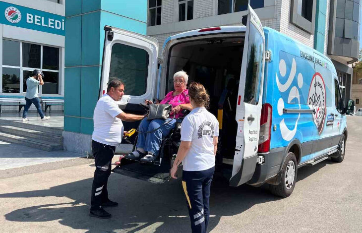 Akülü tekerlekli sandalye kullanıcısı engelli birey, bir minibüsün arkasına özel olarak yapılmış engelli asansörü ile araçtan aküllü sandalye üzerinde indiriliyor. Engelli bireyin yanında refakat ile belediye personelleri var.
