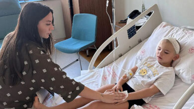 Fotoğrafta, hasta yatağında yatan bir çocuk var. Çocuğun kolunda damar yolu açık ve çocuğun kafasının bir kısmı ile sağ kulağı sargı ile kapatılmış. Çocuğun başında genç bir kadın var ve çocuğun elini tutuyor.