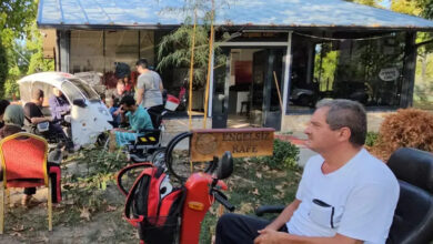 Fotoğrafta, akülü tekerlekli sandalye veya üç tekerli engelli aracı kullanan engelli bireyler bir kafenin önünde oturuyor. Kafenin önünde yer alan yeşil alanda yere tahta bir tabela ile “Engelsiz Kafe” yazısı yazılmış.