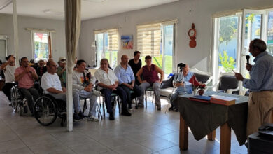 Fotoğrafta, farklı yeti yitimine sahip engelli bireyler bir salonda bir araya gelmişler. Tekerlekli sandalye veya tahta sandalyeler, koltuklar üzerinde oturan engelli bireyler sahnedeki kişiyi dinliyorlar.