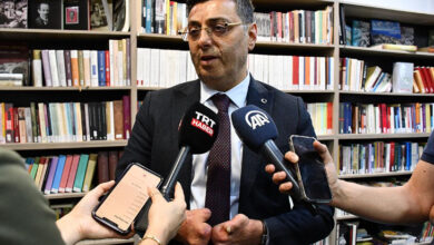 Milletvekili Serkan Bayram, arkasında kitaplıklara dizilmiş kitaplar ve önünde TRT ile AA'nın mikrofonuna basın açıklaması yapıyor.