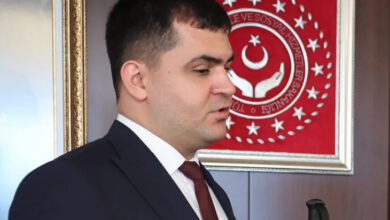 Türkiye'nin ilk engelli hakim yardımcısı olan Sabit Kılıç, Adalet Bakanlığı logosunun önünde ayakta duruyor. Kameraya omuzu dönük bir şekilde fotoğraf çekilmiş.