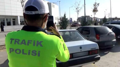 Fotoğrafta, bir otopark alanında trafik polisi sırtında yazan yeşil bir üniforma giymiş erkek otomobillerin fotoğrafını çekerek, hatalı park yapan araçlara ceza yazıyor.
