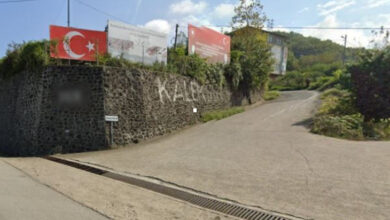 Fotoğrafta, çevresi uzun siyah sayılabilecek taşlarla çevrilmiş bir arazi, arazinin karşısında ise yeşil bir alan ve bir sokağın görüntüsü yer alıyor.