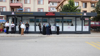 Fotoğrafta yer alan kapalı otobüs durağı önünde bekleyen kalabalık bir insan grubu var. Kapalı otobüs durağının önünde tekerlekli sandalyelerin girebileceği rampa düzeneği yok.