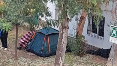 Fotoğrafta bir binanın kenarında ve ağaçların arasında çadır var. Çadır kurulu bir şekilde duruyor. Çadırın dışında ise bir kişinin ayakları gözüküyor.