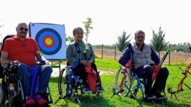 3 tekerlekli sandalyede engelli bireyler oturuyor. 3 engelli bireyden ortada duran kadın, diğerleri erkek sporcular. Engelli sporcuların ellerinde yay ve oklar var. Arkalarında ise okların atıldığı hedef tahtası var.