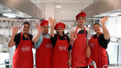 Engelli bireyler ve eğitmen aşçılar tarafından kırmızı renkli mutfak önlüğü giyilmiş. Mutfakta bulunan engelli bireyler ve eğitmen aşçılar kameraya doğru el sallıyorlar.
