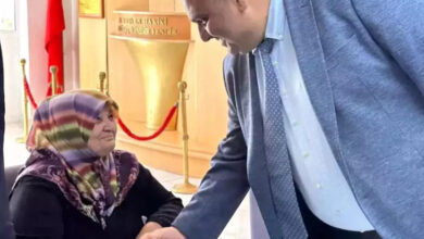 Fotoğrafta, yaşlı bir kadın tekerlekli sandalye üzerinde oturuyor ve karşısında orta yaşlarda bir erkek gülümseyerek elini sıkarak selamlıyor.