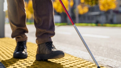 Beyaz Baston ile hareket eden görme engelli bireyin sokakta yönünü bulması için kullanılan sarı renkli çizgiler üzerinde yönünü bulmaya çalışıyor. Görme engelli bireyin sadece belinden aşağısı fotoğrafta yer alıyor.