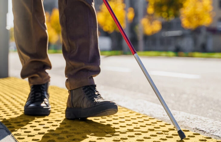Beyaz Baston ile hareket eden görme engelli bireyin sokakta yönünü bulması için kullanılan sarı renkli çizgiler üzerinde yönünü bulmaya çalışıyor. Görme engelli bireyin sadece belinden aşağısı fotoğrafta yer alıyor.