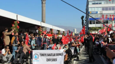 Tekerlekli sandalye kullanan engelli bireyler, bir dar sokaktan kortej ile birlikte ilerlerken en önde yer alan engelli bireyler “Türkiye Sakatlar Derneği İzmir Şubesi” yazılı bir pankart açıyor. Ellerinde Türk Bayrağı olan engelli bireyler sokak boyunca geçiş yapıyor.
